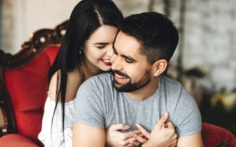 A couple shares a warm, intimate moment. A woman with long dark hair hugs a smiling man from behind as he sits in a red chair. The man has dark hair and a beard, and is wearing a gray shirt. Both are smiling and appear happy.