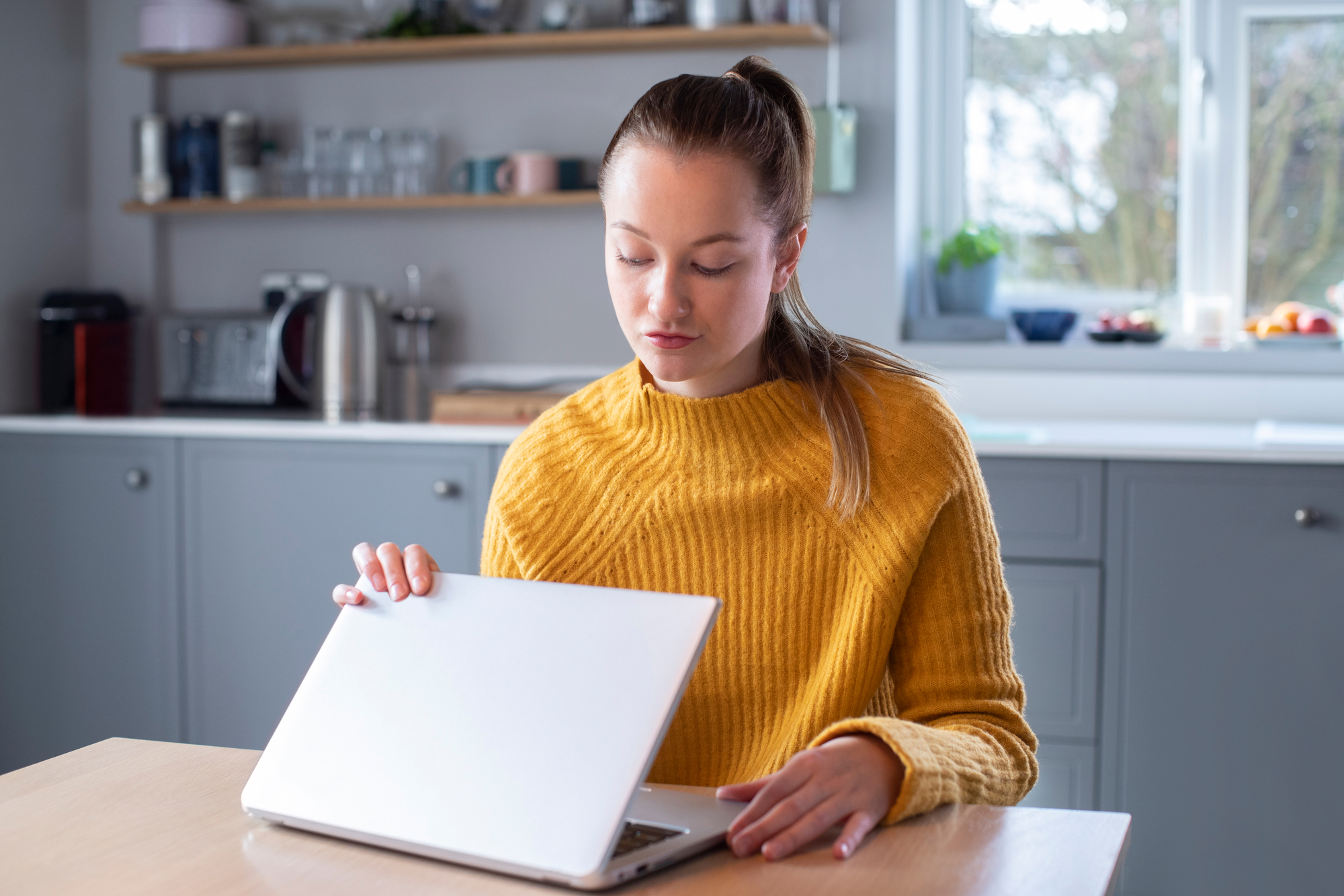 A young woman wearing a yellow sweater is closing her laptop while sitting at a kitchen table. The modern kitchen in the background features gray cabinets, a window, and shelves with various kitchen items. The scene is lit by natural light.