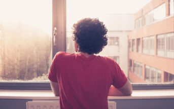 A person with curly hair, wearing a red shirt, is looking out of a window. The outside view shows a modern building with many windows and a slight overcast sky. The lighting suggests it's either early morning or late afternoon.