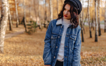 A young woman stands in a sunlit park with autumn trees, wearing a denim jacket, white turtleneck, black hat, and red lipstick. She looks to her left with a serene expression, surrounded by fallen leaves on the ground.