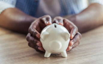 Person's hands carefully holding a white ceramic piggy bank on a wooden table. The background is slightly blurred, focusing attention on the piggy bank. The image conveys themes of saving, finances, or financial security.