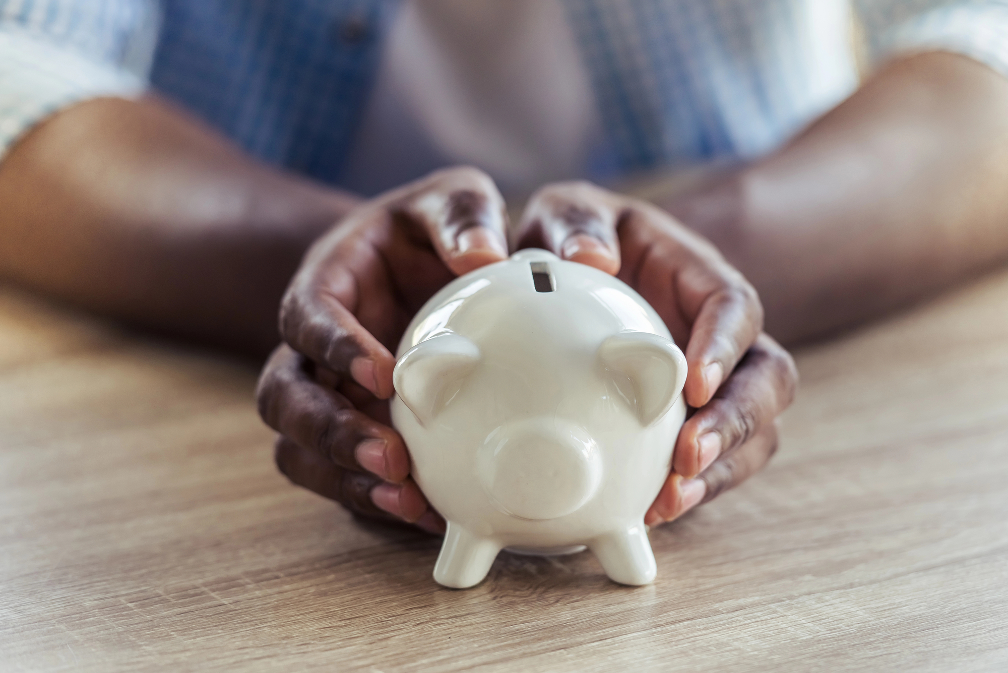 Person's hands carefully holding a white ceramic piggy bank on a wooden table. The background is slightly blurred, focusing attention on the piggy bank. The image conveys themes of saving, finances, or financial security.