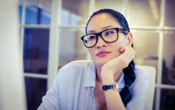 A woman wearing glasses and a light blue shirt is sitting with her chin resting on her hand, appearing thoughtful. She has long, dark hair pulled back in a braid and is positioned in an office or workspace with large windows in the background.