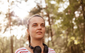 A person with a thoughtful expression stands outdoors in a sunlit forest wearing a red and white striped shirt and black headphones around their neck. Trees and sunlight filtering through the leaves create a serene background.