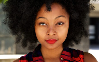 A person with natural curly hair is looking directly at the camera with a calm expression. They are wearing a red and black checkered shirt and have vibrant red lipstick. The background is slightly blurred, indicating an outdoor setting.