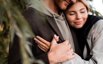 A close-up of a couple embracing. The woman, with a content smile, has her eyes closed and is hugging the man tightly. The man's face is partially visible, and he is wearing a gray hoodie and a brown jacket. Both seem to be outdoors, surrounded by greenery.