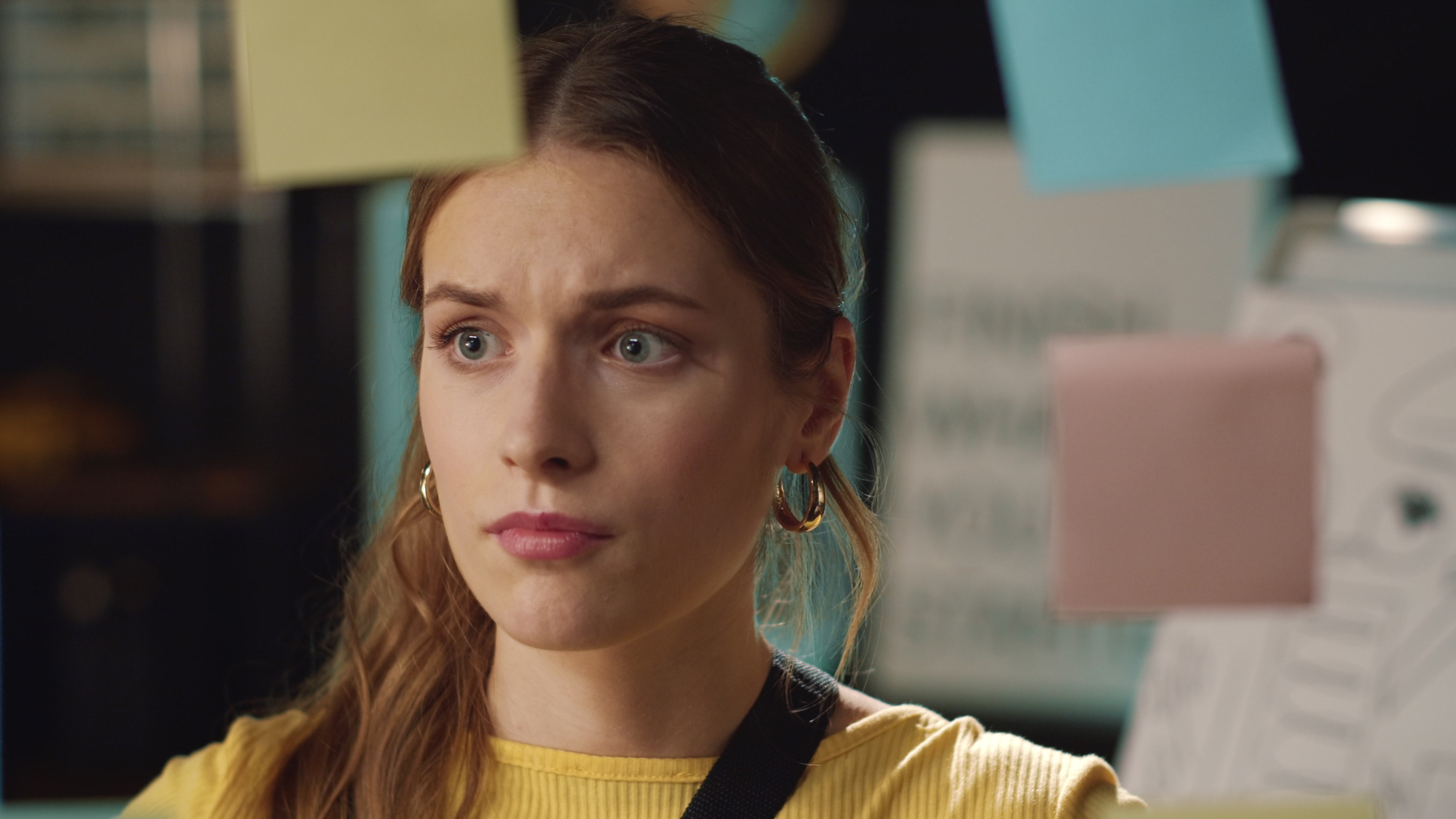 A young woman with long hair and gold hoop earrings looks concerned as she glances at colorful sticky notes in front of her. She is wearing a yellow top, and the background is blurred, suggesting an office or workspace setting.