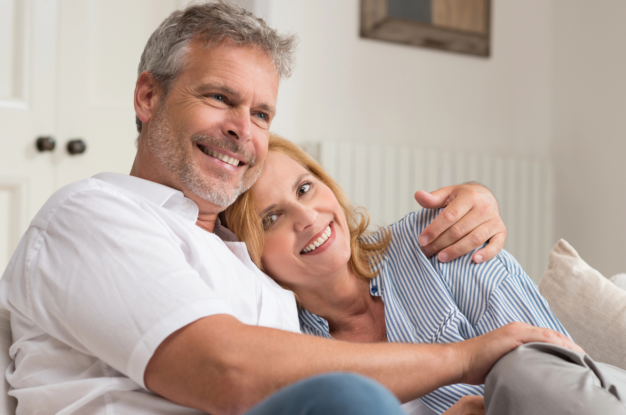 A smiling couple is sitting on a couch, with the man wearing a white shirt and the woman in a blue striped blouse. The man has his arm around the woman as she leans her head on his shoulder. They both appear relaxed and happy in a cozy, light-colored room.