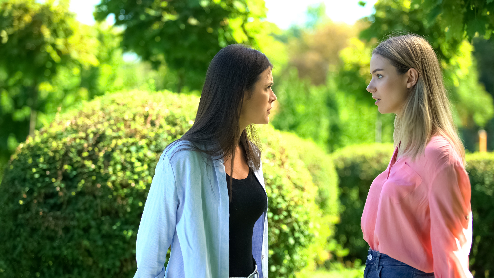 Two women stand outdoors in a park with lush greenery. One woman, in a blue shirt over a black top, appears to be talking to the other woman in a pink shirt. Both have serious expressions, gazing at each other with a background of neatly trimmed bushes and trees.