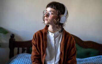 A person with glasses and headphones sits on a bed with a thoughtful expression. They are wearing a white shirt and a brown cardigan. The bed has a wooden headboard and is partially covered by blue-striped bedding. The background is a plain wall.