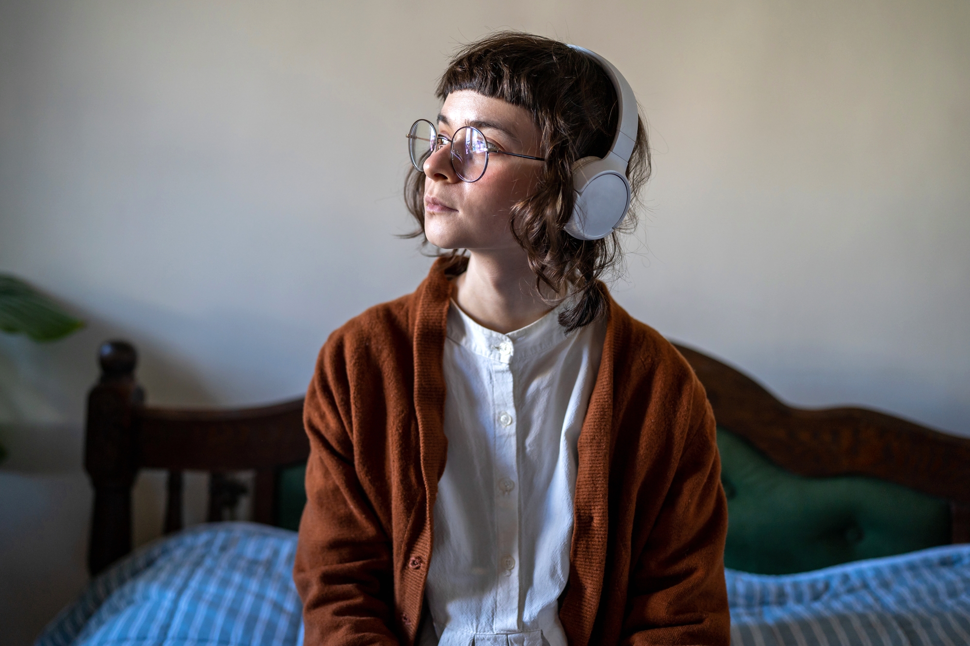 A person with glasses and headphones sits on a bed with a thoughtful expression. They are wearing a white shirt and a brown cardigan. The bed has a wooden headboard and is partially covered by blue-striped bedding. The background is a plain wall.