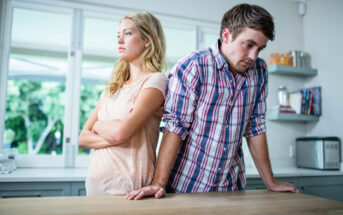 A man and a woman stand in a kitchen. The woman has her arms crossed and looks away, while the man leans on the counter and looks down. The room has modern decor with a window in the background. Both appear to be upset or in disagreement.