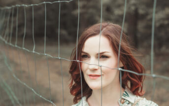 A woman with shoulder-length reddish-brown hair stands behind a wire fence. She wears a light-colored blouse with a subtle pattern and looks slightly to her left with a calm expression. The background is blurry, showing trees and foliage.