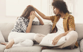 Two women sit on a couch; one covers her face with her hands, appearing upset, while the other gently places a hand on her head, offering comfort. Both wear casual clothes and the room is softly lit, suggesting a supportive and empathetic environment.