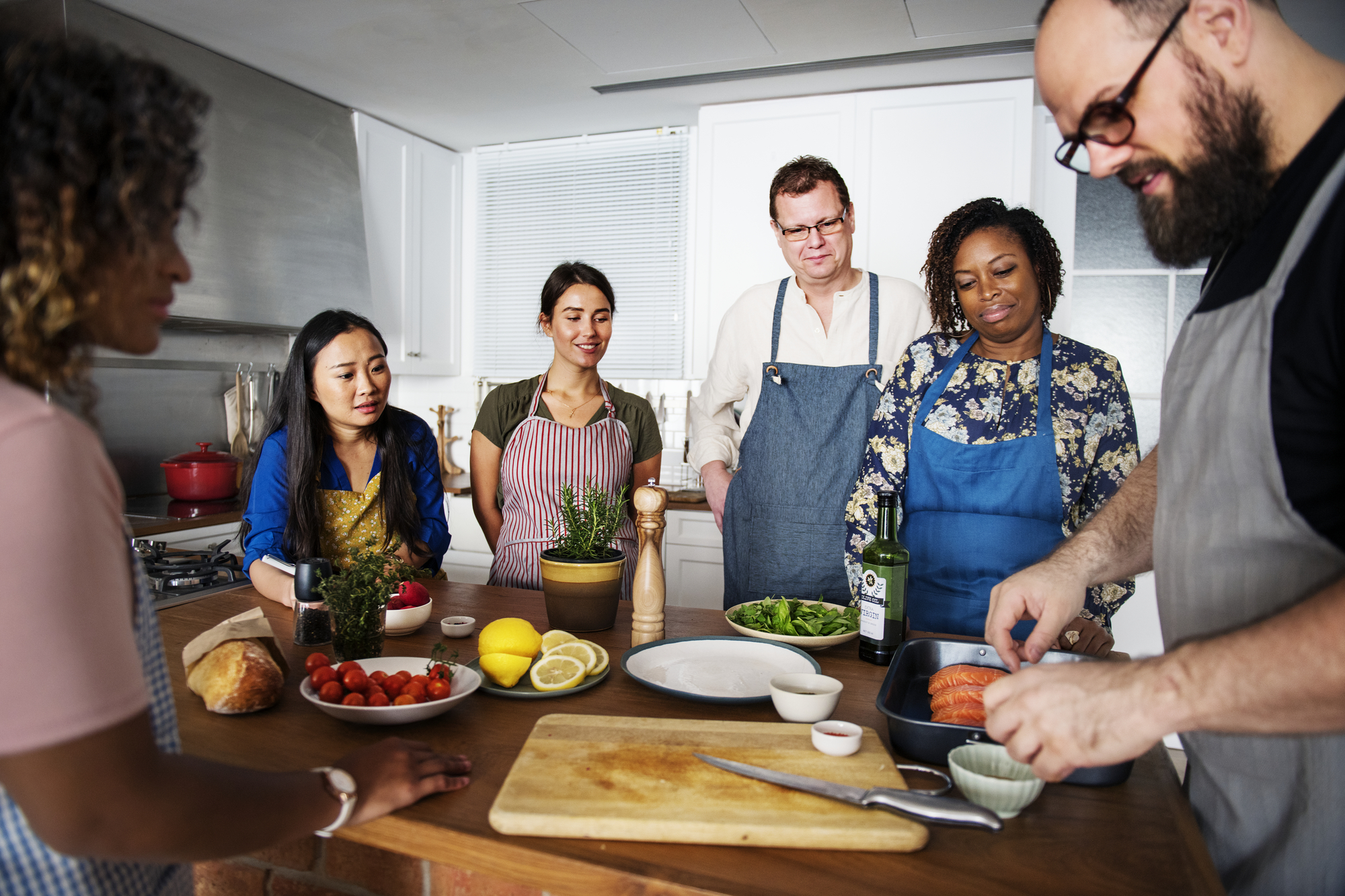 A group of five people gathered around a kitchen island, attentively listening to a man in an apron demonstrating how to prepare salmon. Ingredients such as tomatoes, lemons, and leafy greens, along with a bottle of oil, are spread out on the table.