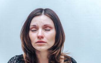 A woman with long brown hair is pictured with her eyes closed, and tears are visible on her face. She appears to be emotional or in distress. She is wearing a black, lacy top, and the background is plain and light-colored.