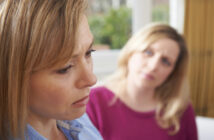 Close-up of two women indoors. The woman in the foreground has a downcast expression and appears upset, while the woman in the background looks at her with a concerned expression. The background is softly blurred, with hints of greenery and a window visible.