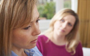 Close-up of two women indoors. The woman in the foreground has a downcast expression and appears upset, while the woman in the background looks at her with a concerned expression. The background is softly blurred, with hints of greenery and a window visible.