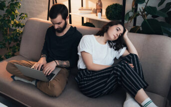 A man with a laptop sits on a couch, focused on typing. A woman next to him, looking distressed, rests her head on her hand. The room has plants and shelves in the background, creating a cozy setting. Both appear to be casually dressed.