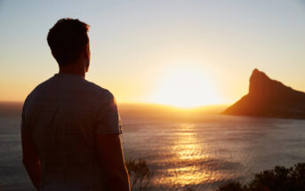 A man in a gray t-shirt stands on a hilltop, gazing at a serene sunset over the ocean. The sun is low on the horizon, casting a warm glow across the water and silhouetting a rugged mountain in the background.