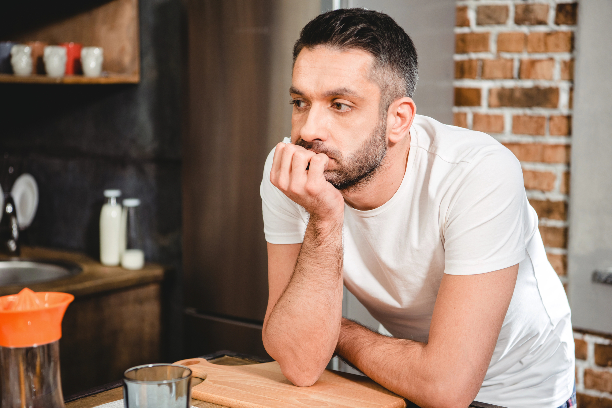 A man with short dark hair, wearing a white t-shirt, leans on a kitchen counter with his chin resting on his hand. He appears deep in thought. The kitchen has a modern yet rustic feel, featuring exposed brickwork and wooden cabinets. Milk and a glass are on the counter.