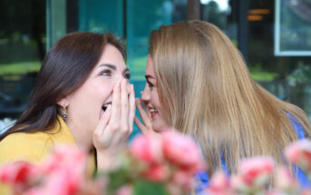 Two women sharing a secret and laughing joyfully. One woman with brown hair holds her hand to her mouth in surprise, while the other woman with blonde hair leans in closely. They are sitting near a table with pink flowers in the foreground.