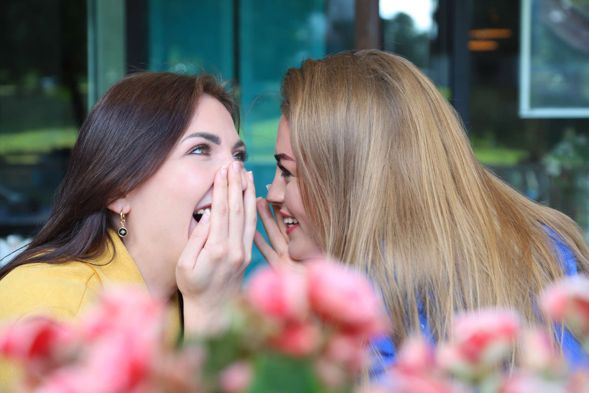 Two women sharing a secret and laughing joyfully. One woman with brown hair holds her hand to her mouth in surprise, while the other woman with blonde hair leans in closely. They are sitting near a table with pink flowers in the foreground.