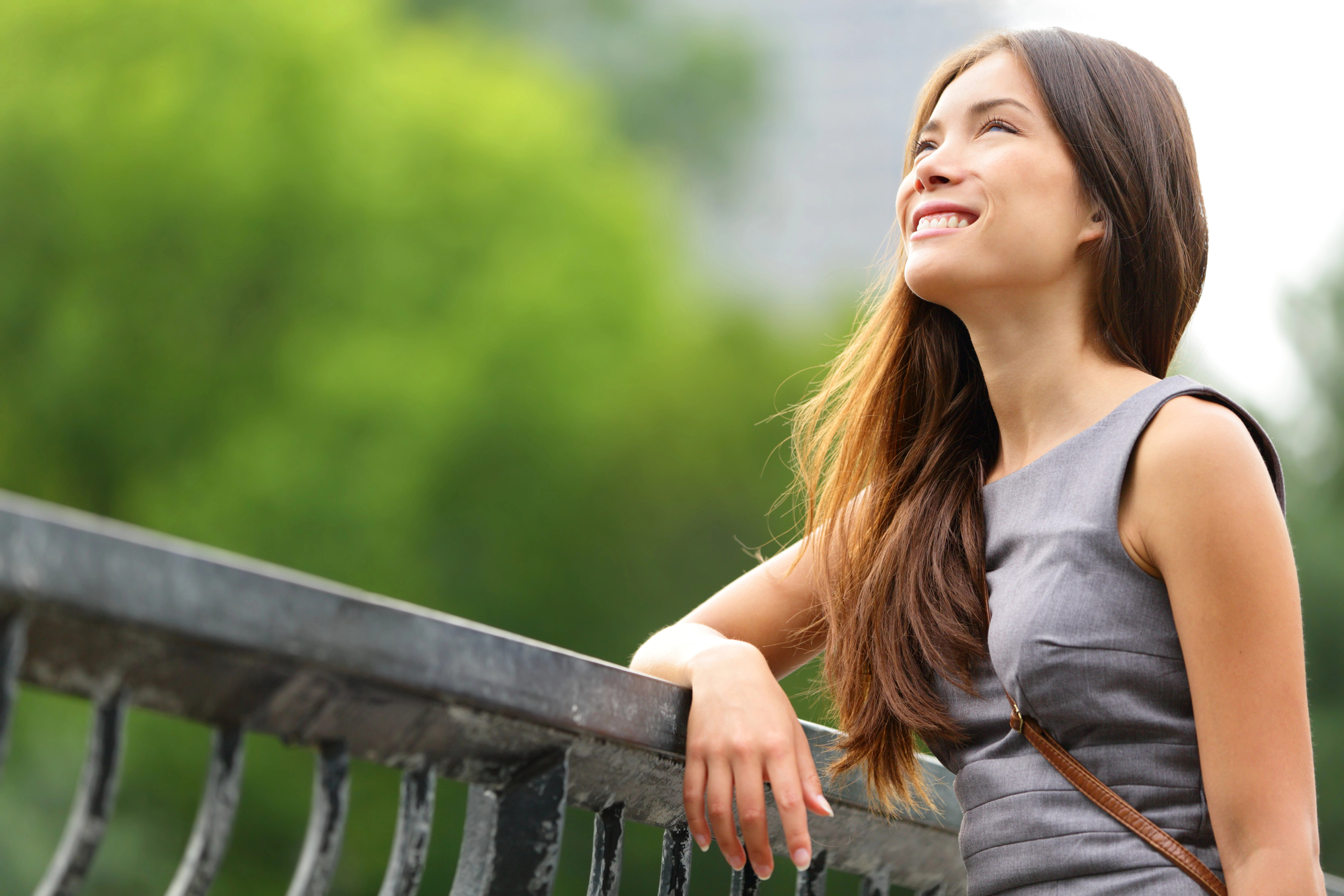 A woman with long hair is leaning on a metal railing outdoors, smiling and looking up. She is wearing a sleeveless gray dress, and there is a green, blurred background suggesting a park or natural setting.