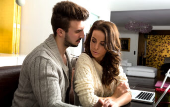 A couple sits closely together on a leather couch at home, both wearing cozy sweaters. The man has a serious expression and gently touches the woman's arm, while she looks down pensively. A laptop is open on the table in front of them, and the room is warmly lit.