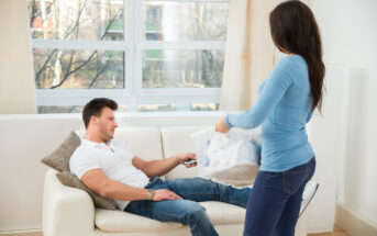 A man is lounging on a sofa with a remote control in his hand, looking at a woman who is standing nearby holding a laundry basket full of clothes. The room features large windows with a view of trees outside.