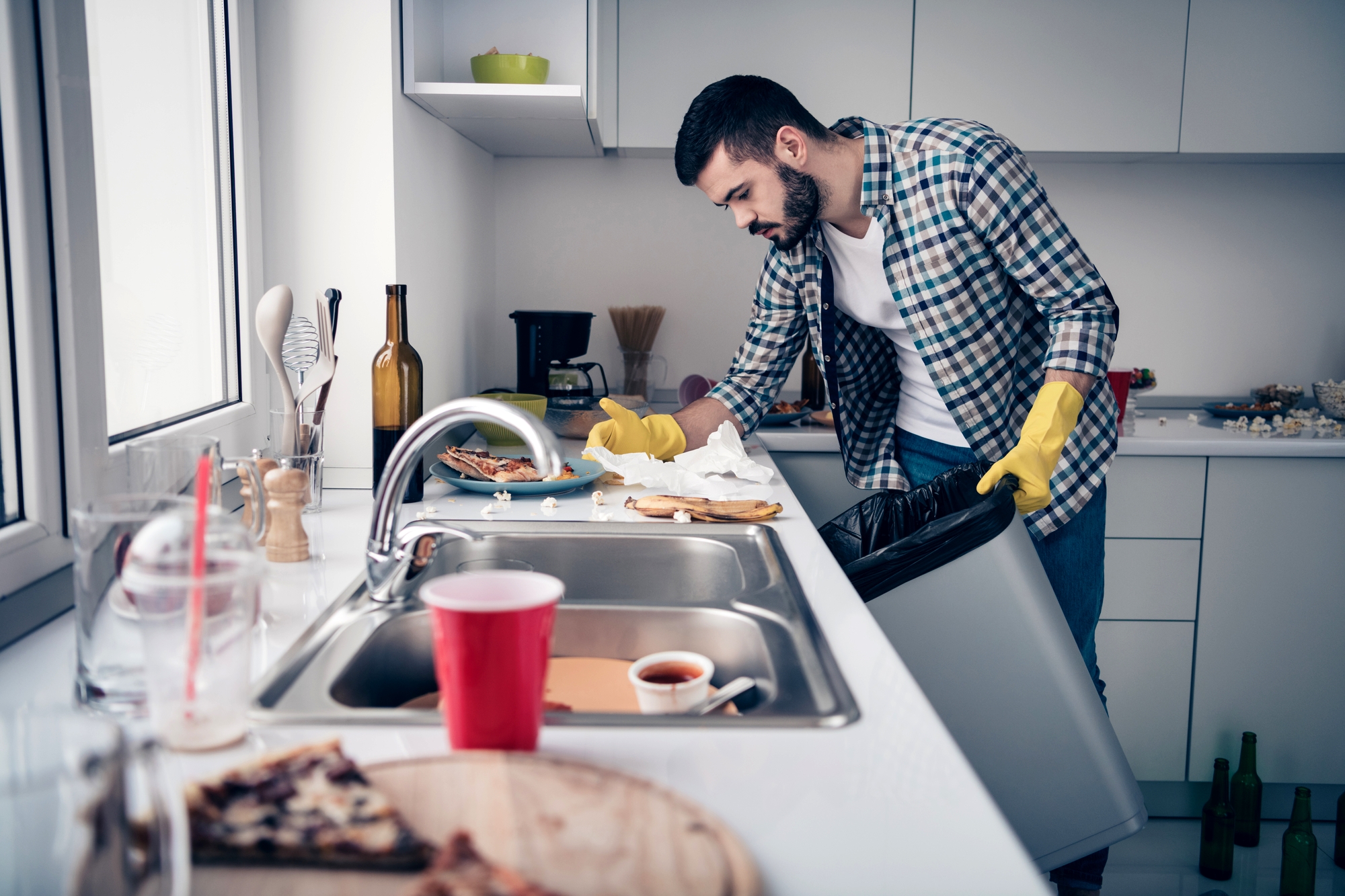 A man wearing yellow gloves tidies up a modern kitchen. He puts food scraps into a trash bag while standing near a sink filled with dishes. The countertop holds leftover pizza, a coffee cup, and various kitchen items. The atmosphere appears busy and cluttered.