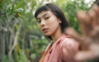 A young woman with dark hair and bangs is standing outdoors, surrounded by greenery. She is wearing an orange top and a red gingham-patterned jacket. She extends one arm towards the camera, slightly out of focus. The expression on her face is neutral.