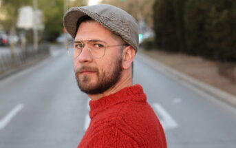 A man with a beard and glasses wearing a red sweater and a gray flat cap looks over his shoulder while standing on an empty road lined with trees. The background is slightly blurred, giving emphasis to his facial expression.