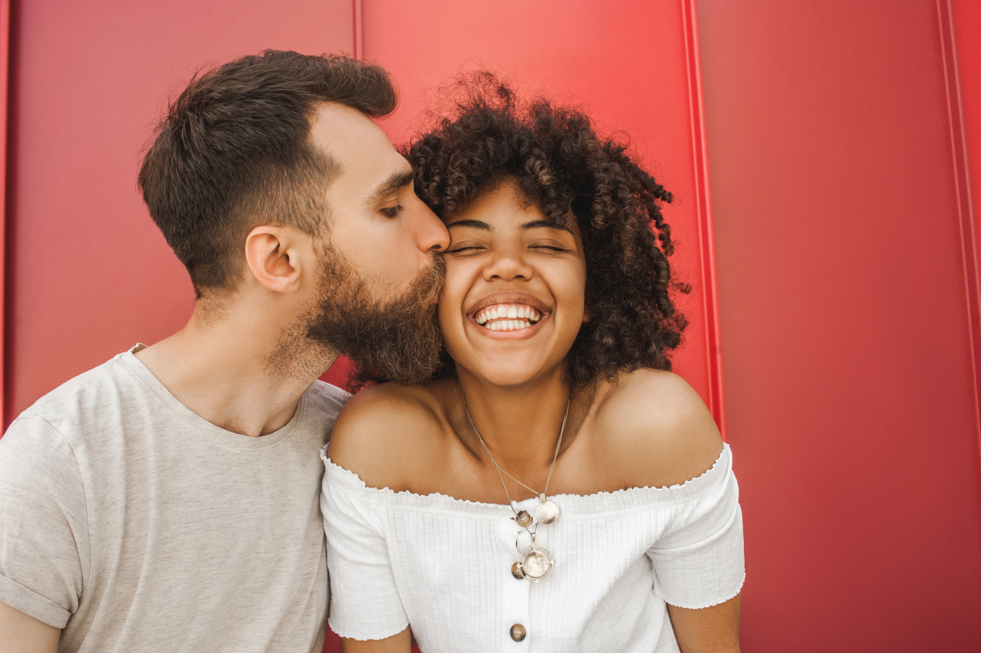 A man with a beard kisses a smiling woman on the cheek. They are in front of a vibrant red background. The woman is wearing an off-shoulder white top and a large seashell necklace. The man is wearing a light gray shirt. Both appear happy and relaxed.