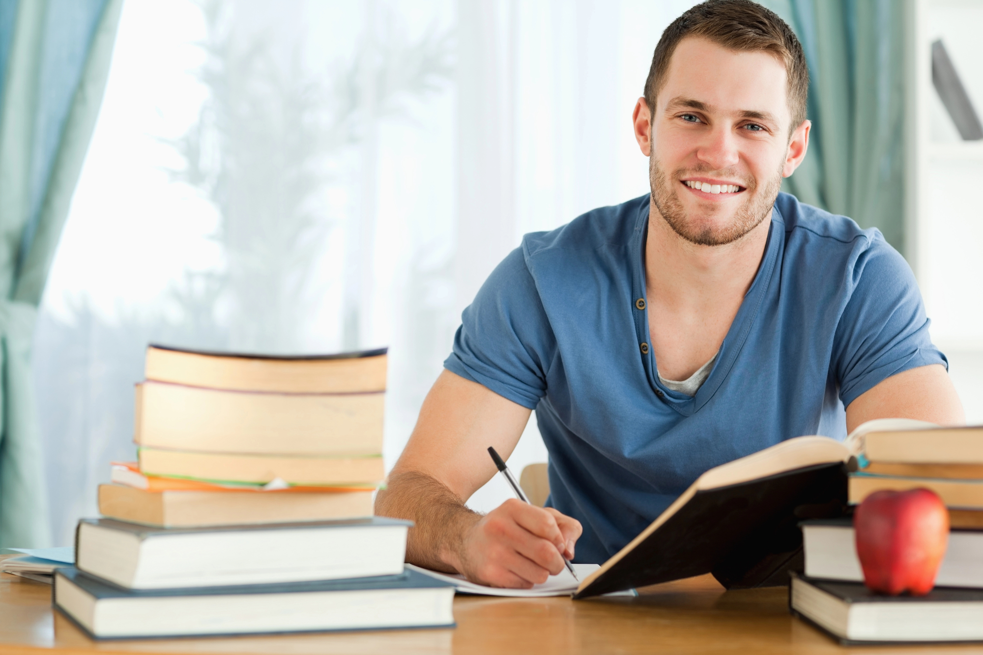 A young man with a beard, wearing a blue t-shirt, sits at a desk with an open book, taking notes. Several stacked books and a red apple are on the desk, and a window with sheer curtains is in the background. He is smiling at the camera.