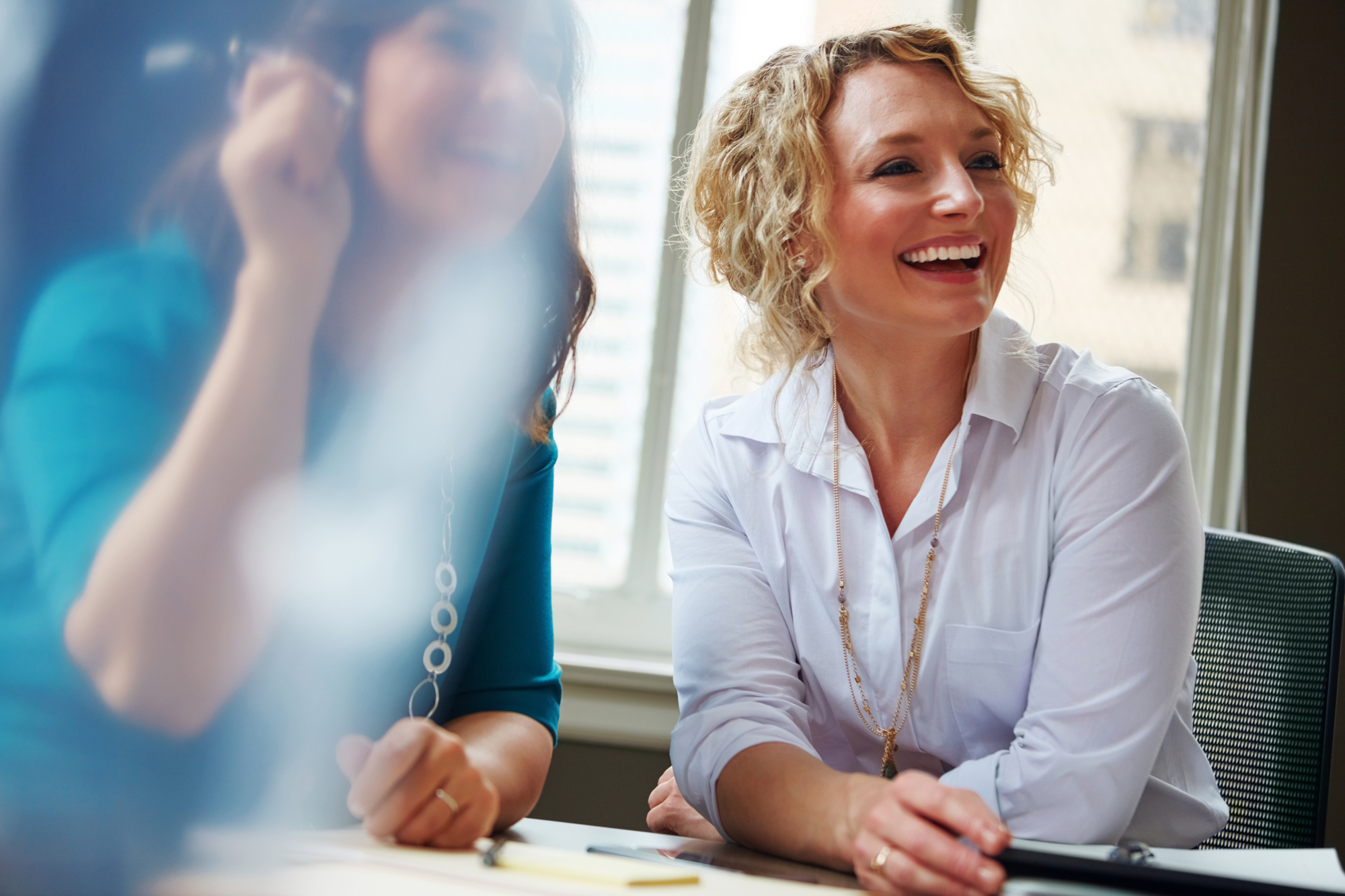 Two women sitting at a table, laughing and smiling. One is wearing a white blouse and has curly blonde hair, while the other is blurred in the background, wearing a turquoise top. They are in a bright room with large windows.