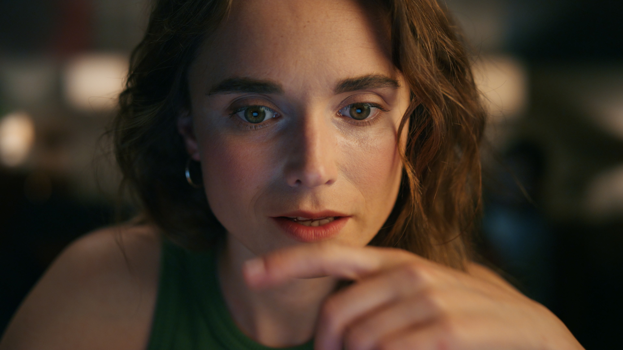 A woman with wavy brown hair and green eyes is intently looking at something off-camera. She is wearing a green sleeveless top and a silver hoop earring. Her hand is raised to about chin level, with her index finger pointing outward. The background is blurred.