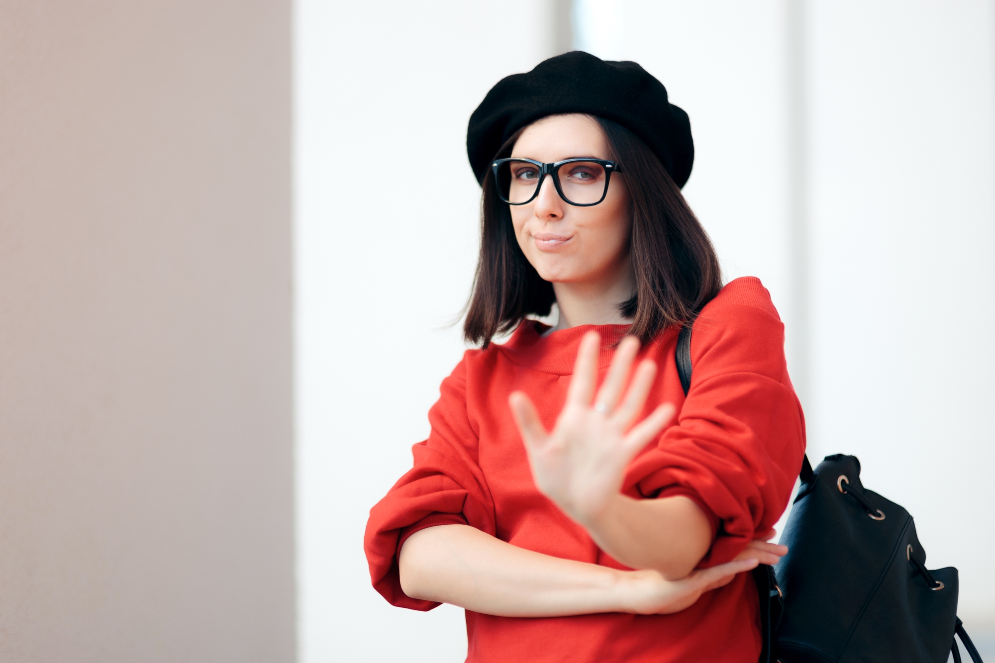 A person with shoulder-length dark hair, wearing a black beret, eyeglasses, and a red sweater, is extending one hand forward in a "stop" gesture while looking directly at the camera. They also have a black backpack over one shoulder.