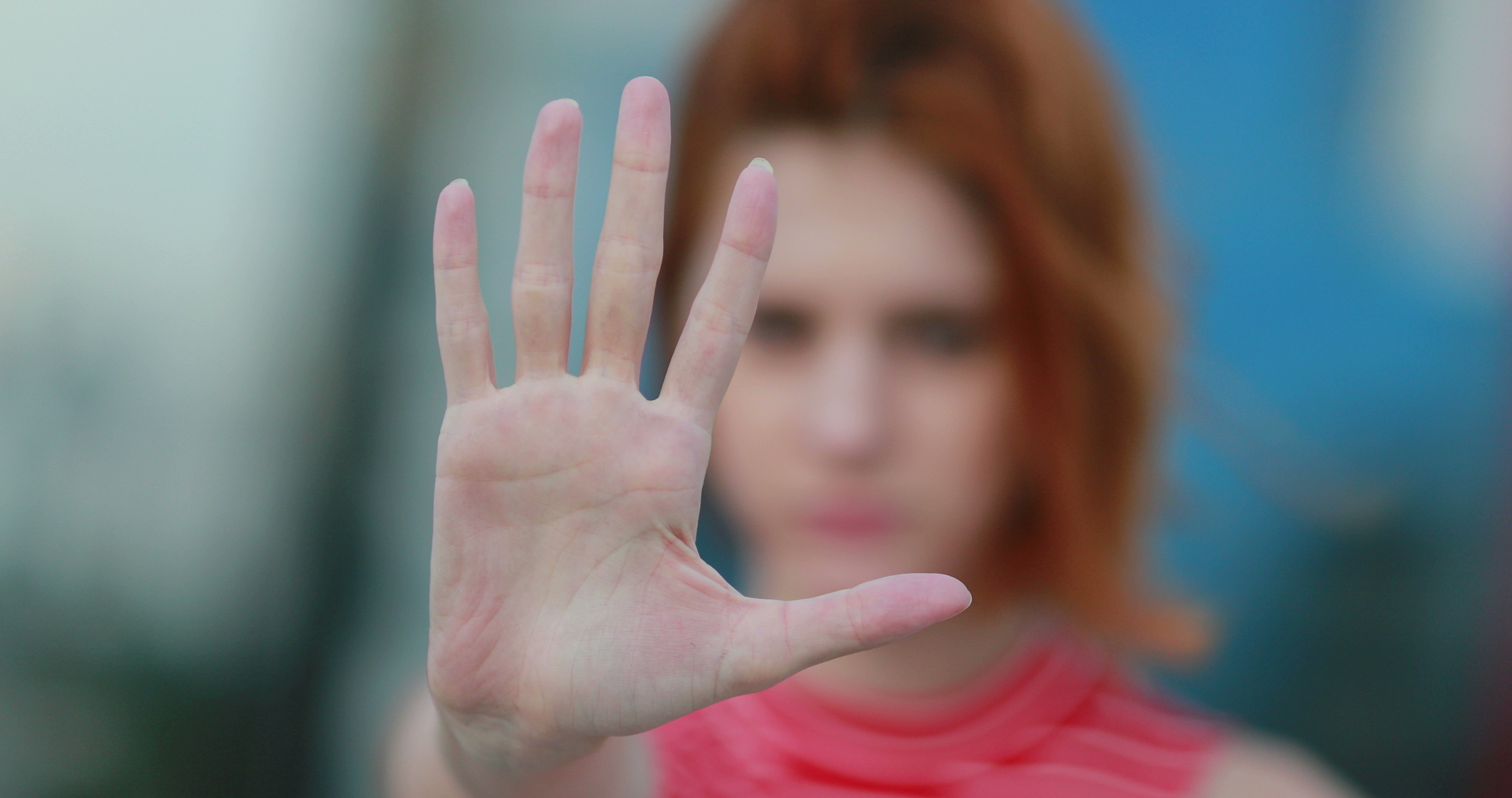 A person with red hair is shown close-up with an outstretched hand facing the camera, fingers spread, in a gesture that could signify stopping or shielding. The background is blurred, directing focus to the hand and the person's vibrant hair.