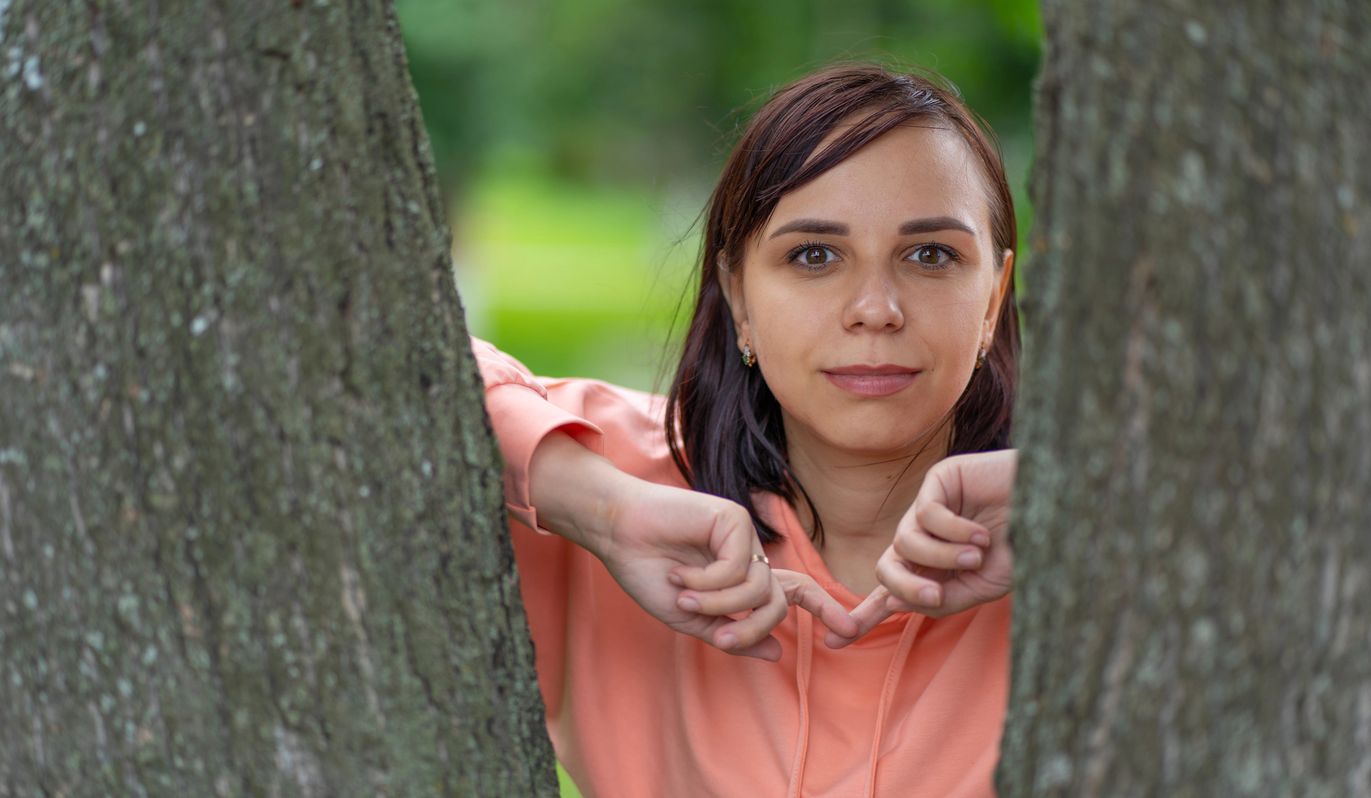 A woman in a peach hoodie looks directly at the camera, framed between two tree trunks. She has short brown hair and a serene expression. Blurred greenery is visible in the background, suggesting a natural outdoor setting.