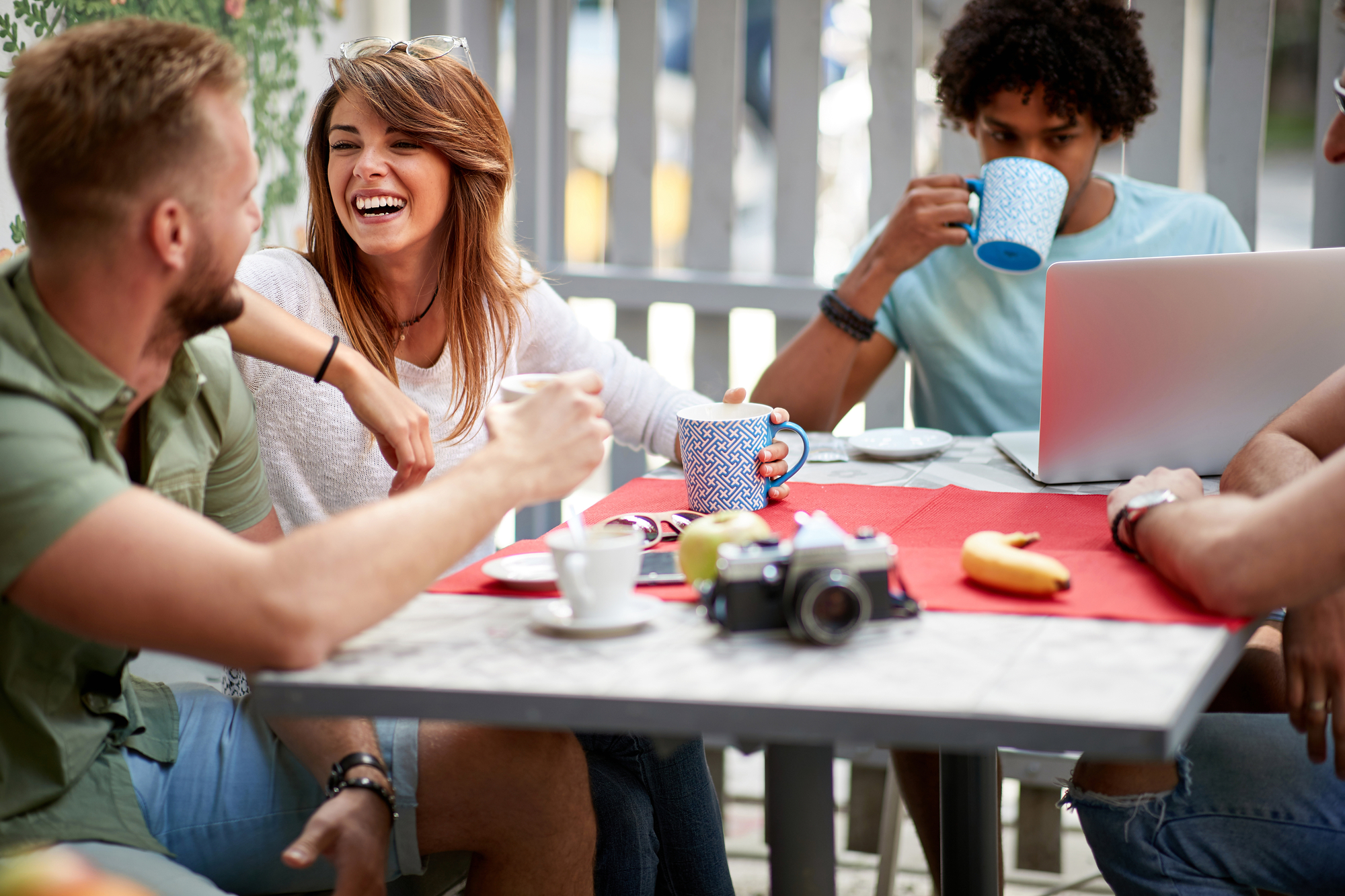 A group of four people sit at an outdoor table, laughing and drinking coffee. A laptop is on the table with a camera and some bananas. The background includes plants and a partially visible street. The scene is casual and cheerful.