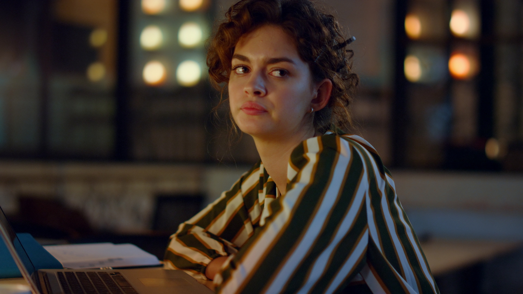 A woman with curly hair, wearing a green and white striped shirt, sits pensively in a dimly lit room in front of a laptop. She has her arms crossed and looks slightly to her left. Blurred lights and a window are visible in the background.