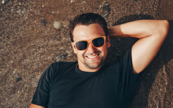 A man with short dark hair is lying on his back on a sandy beach, smiling and wearing orange-framed sunglasses and a black T-shirt. His right arm is bent and resting behind his head. The beach is covered with small pebbles and wet sand.