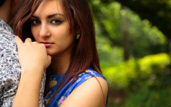 A woman with long brown hair wearing a blue floral dress is standing outdoors. She is resting her hand on her chin and looking directly at the camera. The background is filled with greenery, suggesting a garden or park setting.