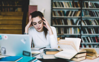 A person with long, dark hair tied back, wearing a white shirt, sits at a desk with an open laptop and several books. They rest their head in their hands, appearing stressed or tired, in a room with shelves filled with books.