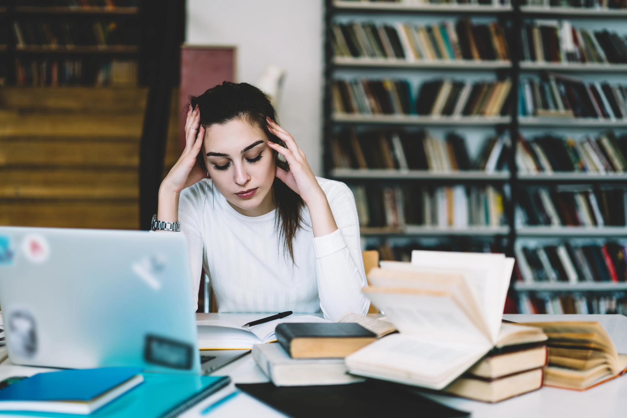 A person with long, dark hair tied back, wearing a white shirt, sits at a desk with an open laptop and several books. They rest their head in their hands, appearing stressed or tired, in a room with shelves filled with books.