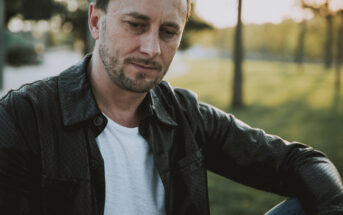 A man in a white t-shirt and dark jacket sits outdoors in a park-like setting, with trees and grass in the background. He is looking down with a thoughtful or contemplative expression. The scene is illuminated by soft, natural sunlight.