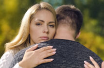 A woman with blonde hair appears serious as she embraces a man whose back is facing the camera. They stand outdoors with a blurred, green natural background.