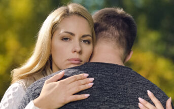 A woman with blonde hair appears serious as she embraces a man whose back is facing the camera. They stand outdoors with a blurred, green natural background.