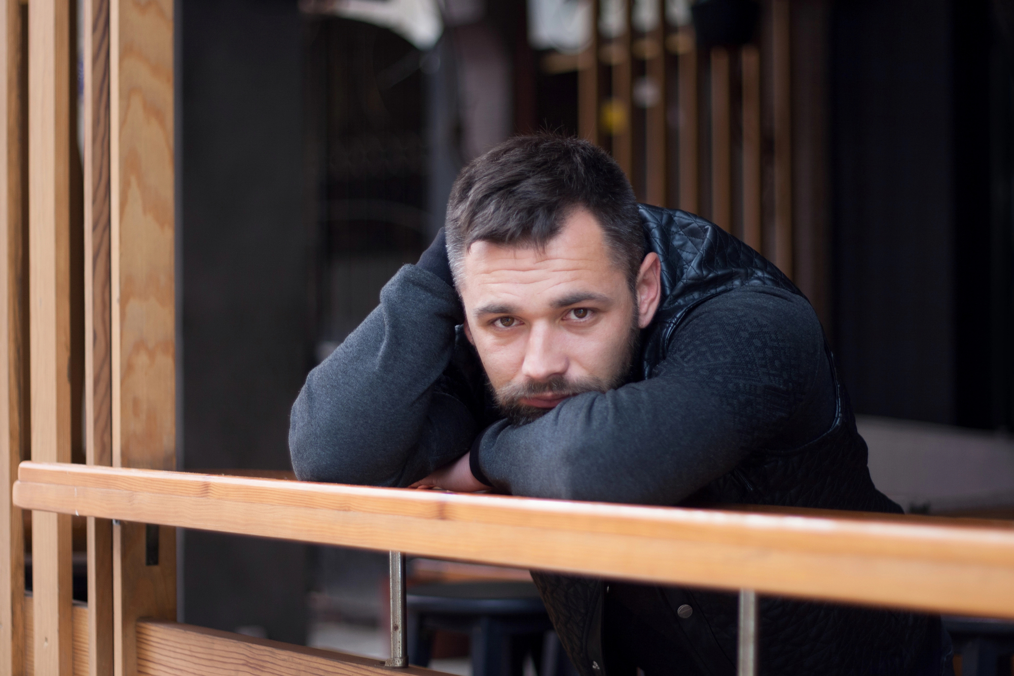 A man with short dark hair and a beard leans on a wooden railing, resting his head on his arms. He is wearing a dark, long-sleeve sweater and a puffy vest. The background is blurred, featuring wooden structures and a partially visible interior space.